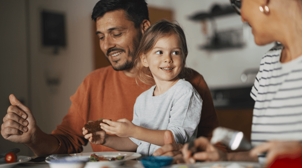 Girl at the dinner table smiling sitting on her fathers lap while looking at her mother.